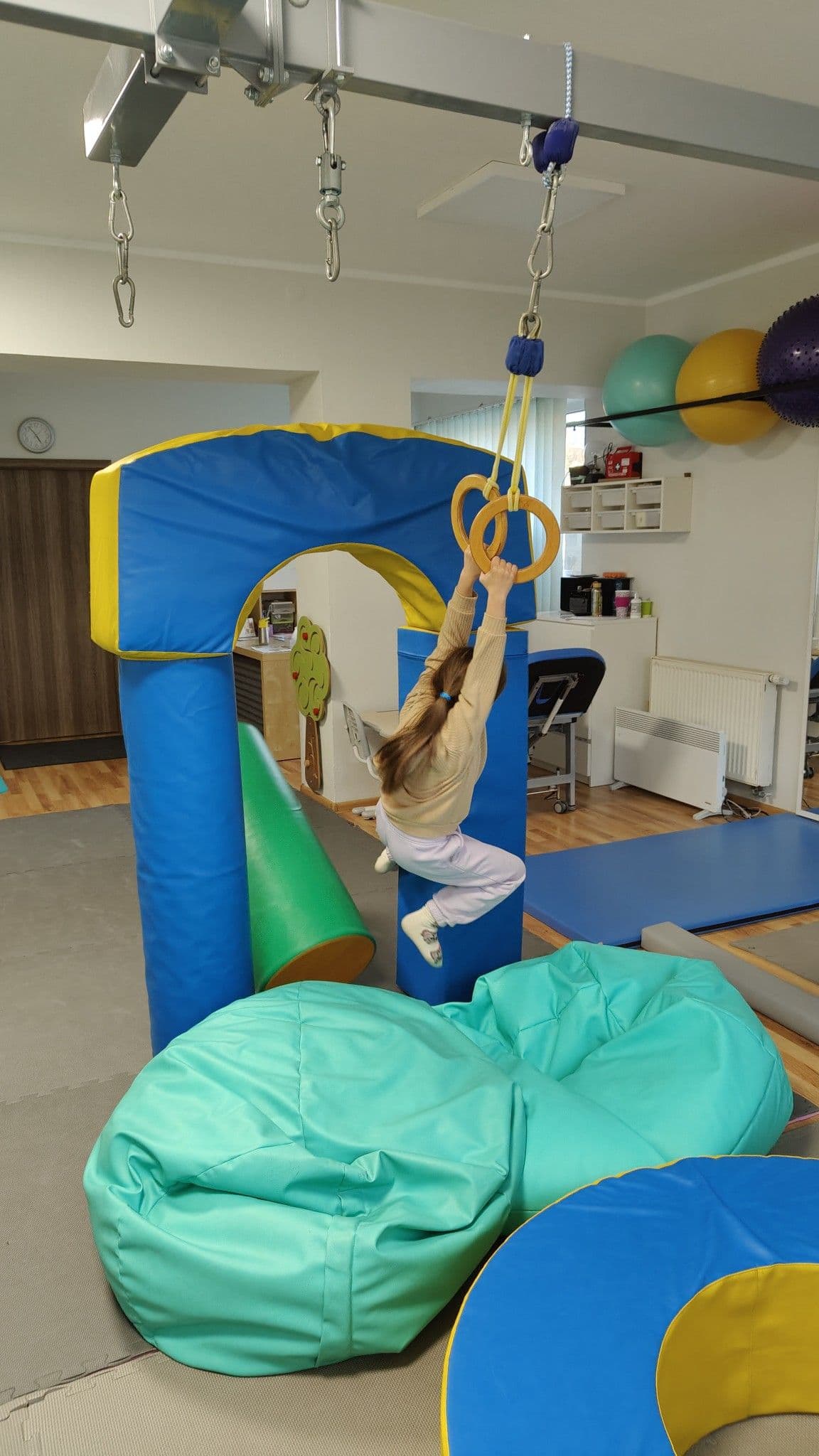 Terapia SI - dziewczynka wisi na obręczach i przewraca nogami klocki piankowe