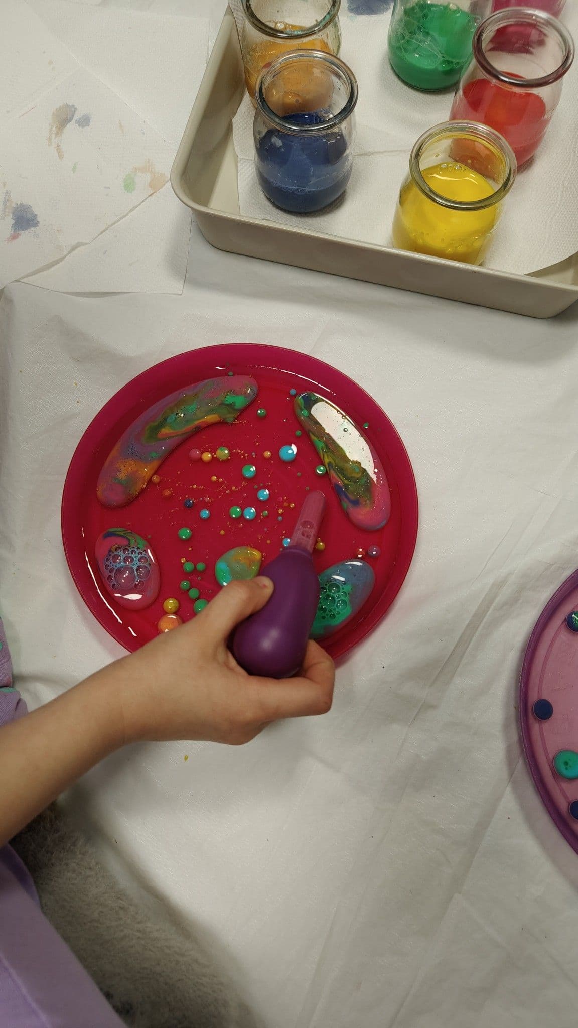 Terapia ręki - zbliżenie na rękę dziecka trzymającego pipetę wypełnioną kolorowym barwnikiem, który wylewa na talerz z olejem